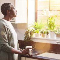 A woman feeling the burden of caregiving gazes wistfully out the window while holding a cup of coffee.