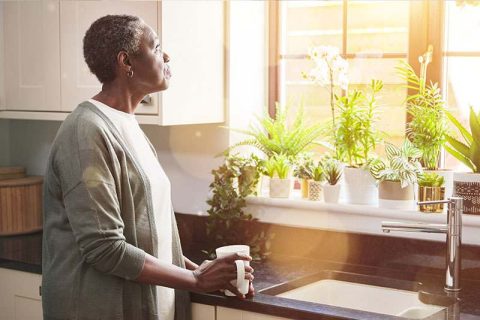 A woman feeling the burden of caregiving gazes wistfully out the window while holding a cup of coffee.