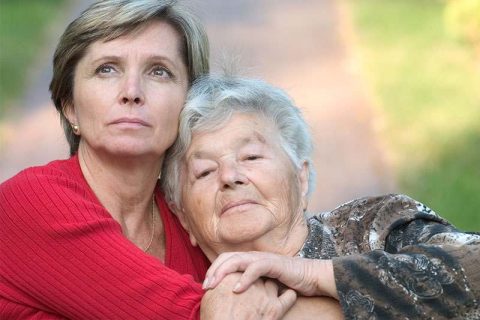 A woman whose mom has dementia gives her a hug while staring thoughtfully into the distance.