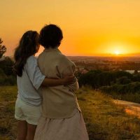 A woman whose family member with dementia becomes anxious at night puts her arm around her as they gaze at the sunset together.