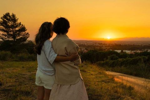 A woman whose family member with dementia becomes anxious at night puts her arm around her as they gaze at the sunset together.