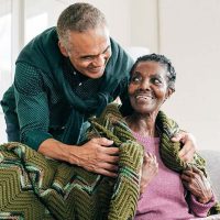 A man utilizing effective Alzheimer’s care tips smiles as he wraps a blanket around his wife’s shoulders.