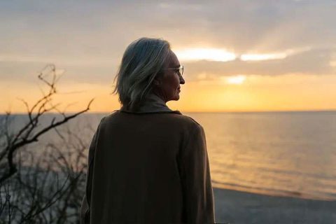 A woman caring for someone later in life gazes out over the ocean at sunset.