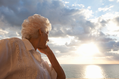 A woman gazes at the sun setting over the water as she struggles with summertime grief for seniors.