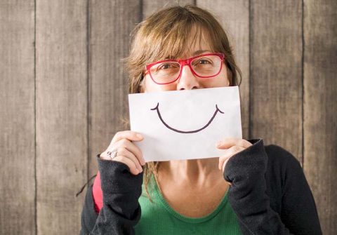 A woman holds up a smiley face mask to indicate that she’s pretending to be fine while caring for a parent with dementia.